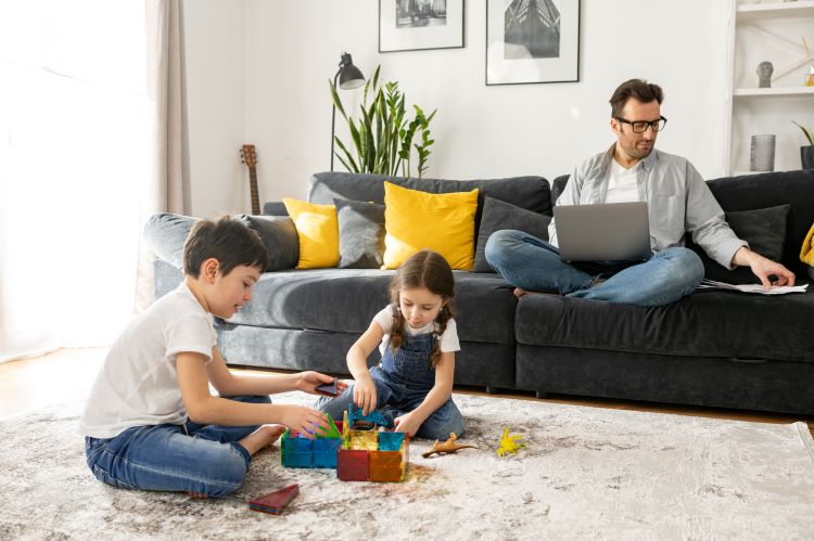 Father using laptop for freelance work sitting on the sofa at home while his two kids playing peacefully together on the floor. Busy single parent is babysitting and working on the distance
